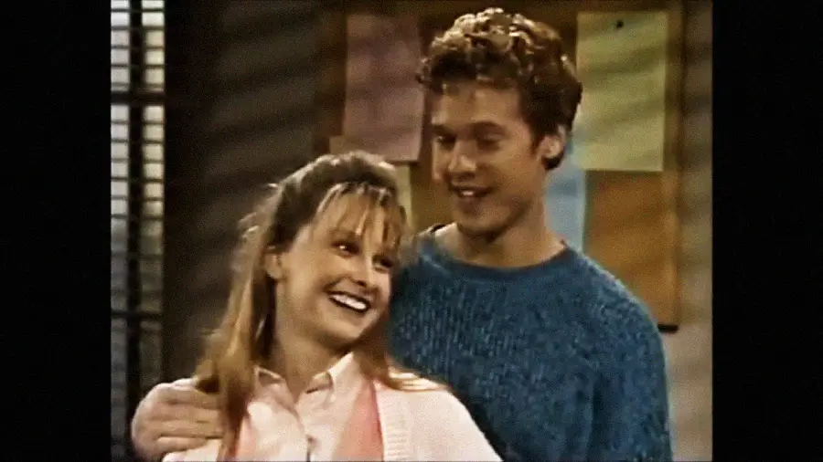 A smiling woman with blonde hair stands in front of a man in a blue sweater, who is smiling and has his arm around her shoulders. They appear happy and are indoors, with a bulletin board in the background.