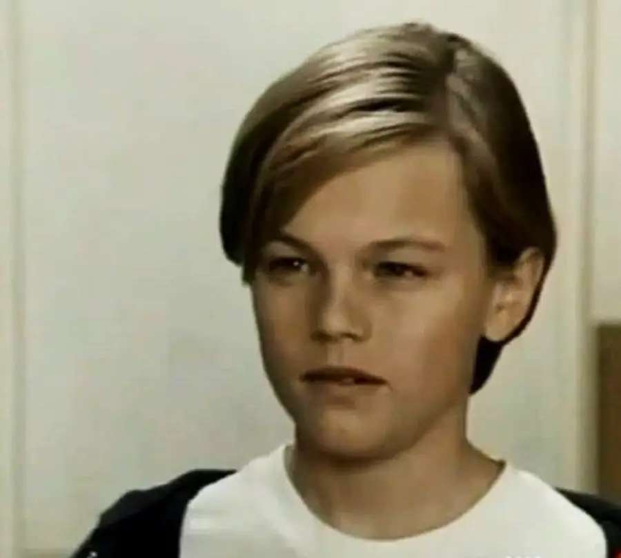 A young boy with straight, light brown hair and a neutral expression, wearing a white shirt and dark jacket, stands indoors against a plain background.