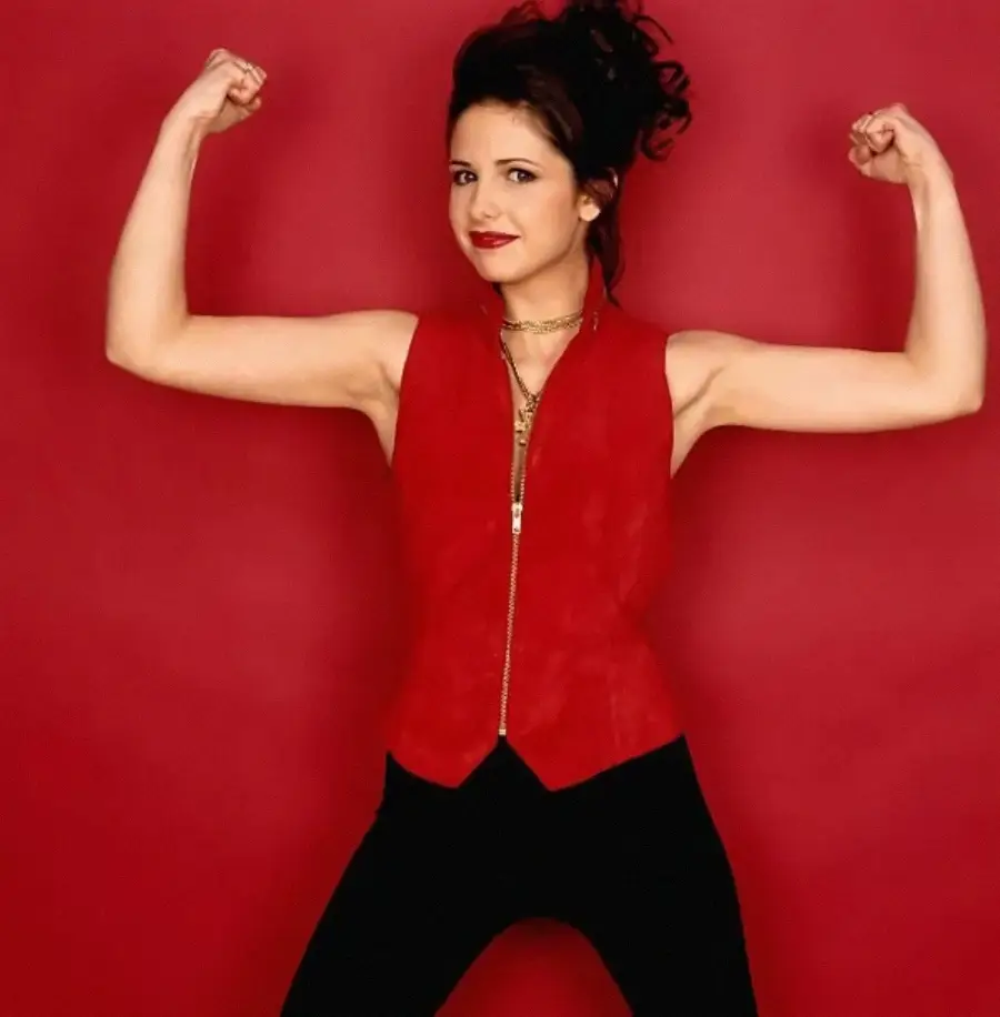A woman stands in front of a red background, flexing her arms and smiling. She wears a red sleeveless vest, black pants, gold necklaces, and has dark hair styled up.