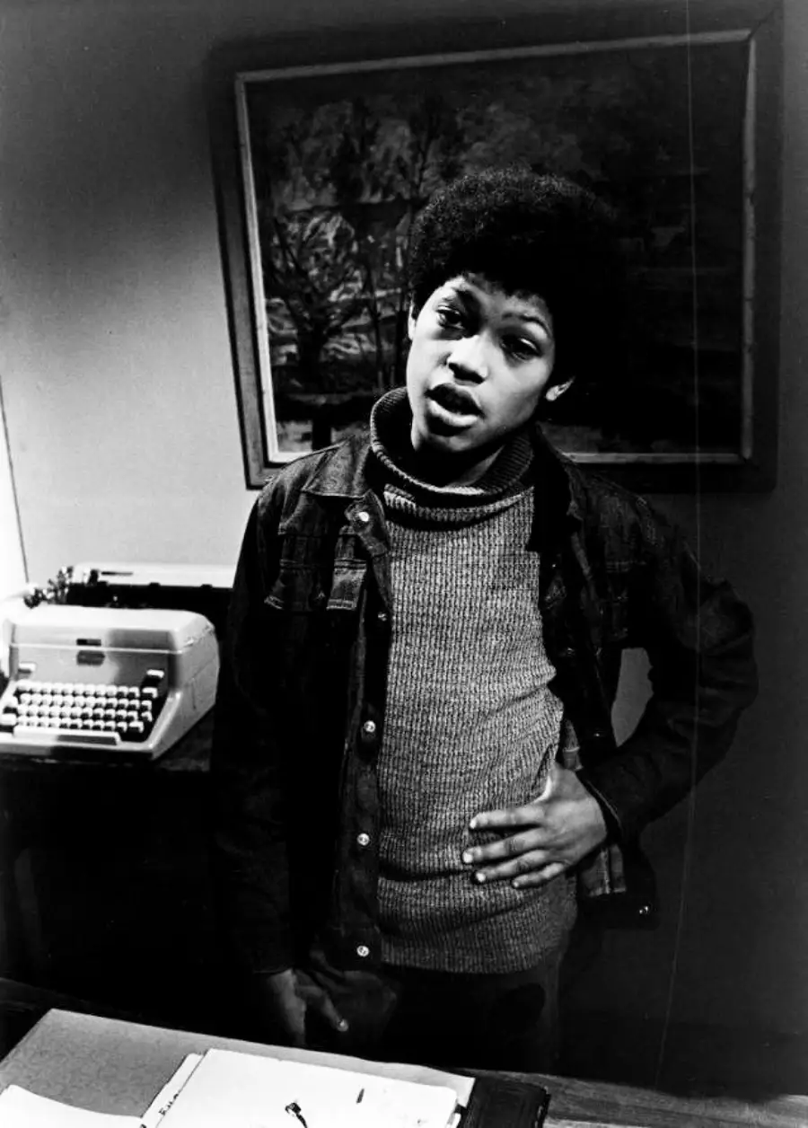Black and white photo of a young person with an afro standing indoors, wearing a jacket and sweater, with one hand on their stomach. A typewriter and painting are visible in the background.