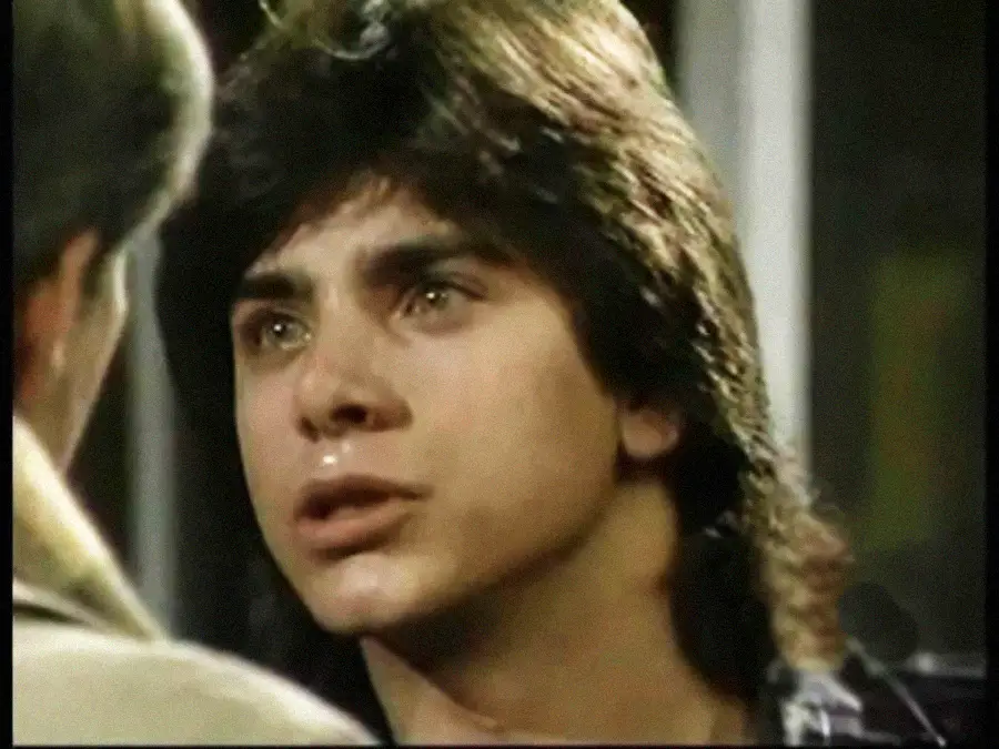 A young man with dark hair and teary eyes looks emotional as he faces another person, who is partially visible from behind.
