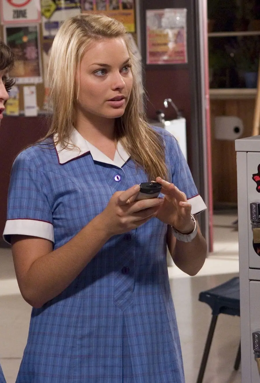 A young woman in a blue plaid school uniform stands indoors, holding a phone with both hands and looking ahead with a focused expression. She has long blonde hair and there are posters on the wall behind her.