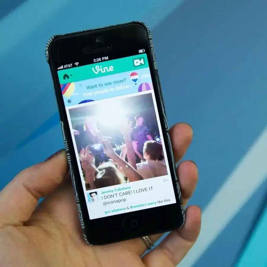 A hand holds a smartphone displaying a Vine video on its screen, featuring a concert scene with people raising their hands and bright stage lights in the background.