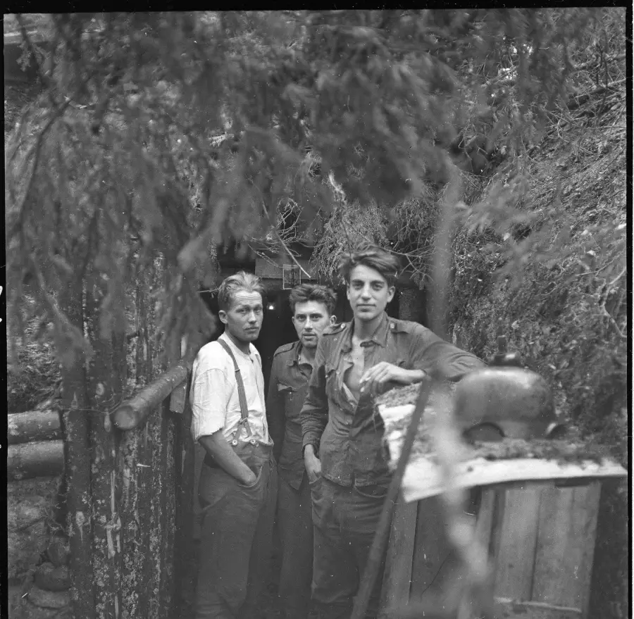 Three young men stand at the entrance of a trench or dugout surrounded by trees and rough wooden structures, looking toward the camera in a black and white outdoor setting.