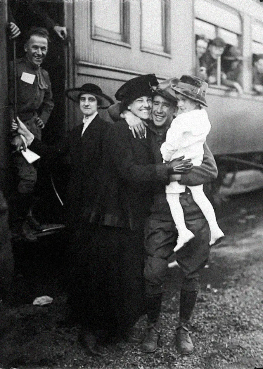 A smiling soldier in uniform hugs a woman and holds a young girl, while another woman stands nearby. Other soldiers and passengers look on from a train beside them. The scene appears joyful and emotional.