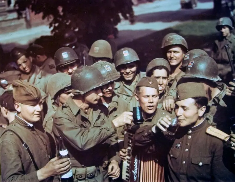 A group of World War II soldiers, both American and Soviet, wearing military uniforms and helmets, stand together smiling and raising drinks in a celebratory toast. One holds an accordion; trees and buildings are in the background.