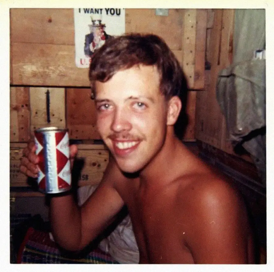 A shirtless young man with a mustache smiles and holds up a can of Budweiser beer. He is indoors, sitting in a wooden-paneled room with an “I Want You” Uncle Sam poster on the wall behind him.