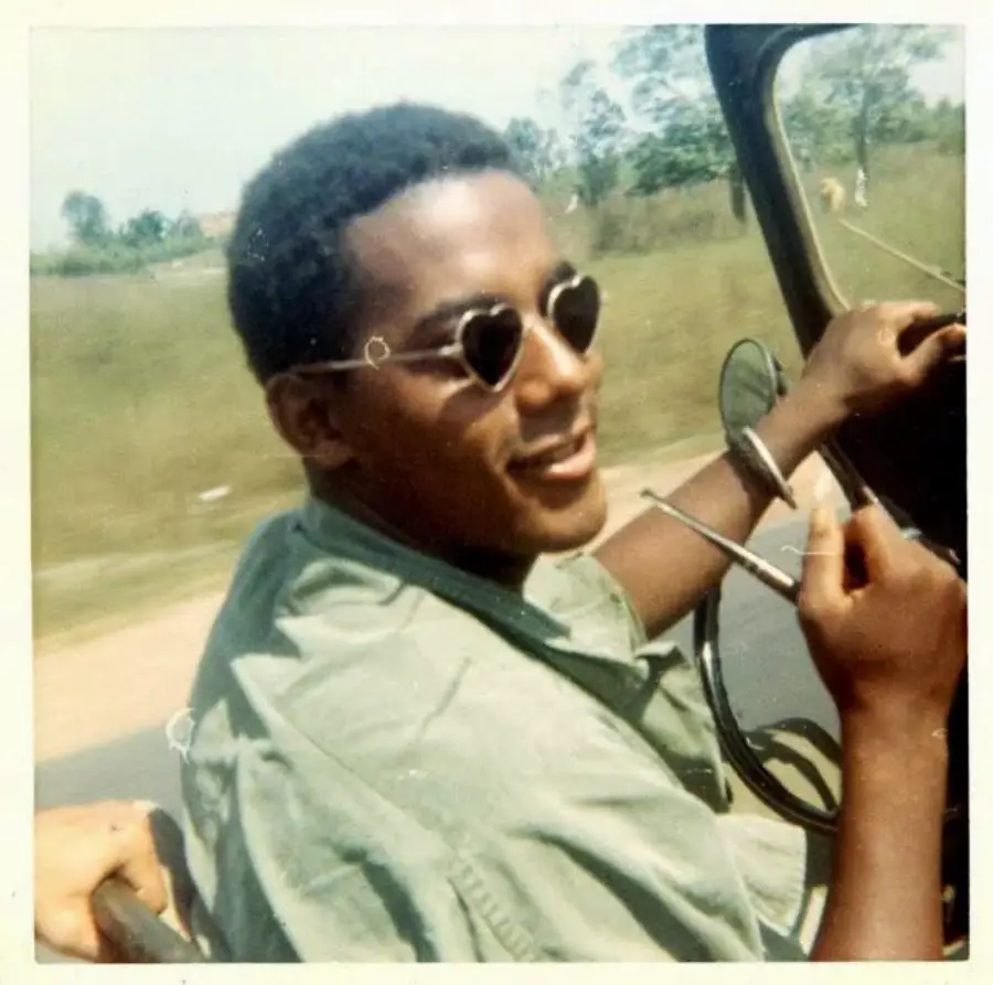 A young man wearing heart-shaped sunglasses smiles while driving a car, holding a cigarette in one hand. The background shows a blurred green landscape.