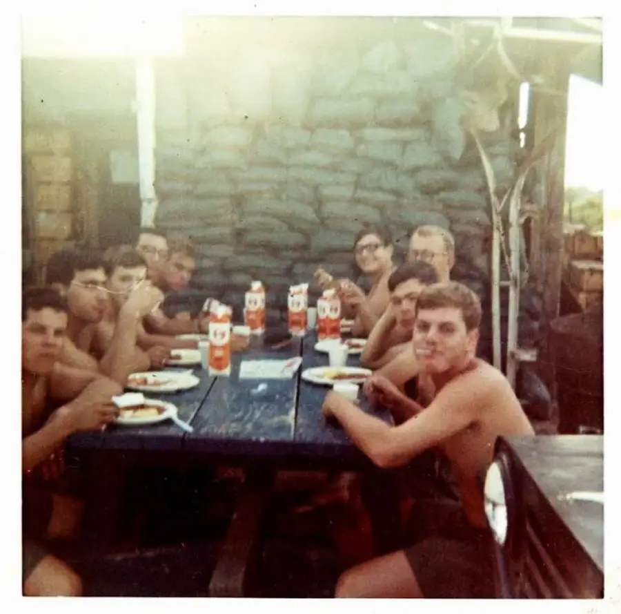 A group of young men sit shirtless around a picnic table outdoors, eating a meal together with plates and orange drink containers. The setting appears rustic, with sandbags and makeshift structures in the background.