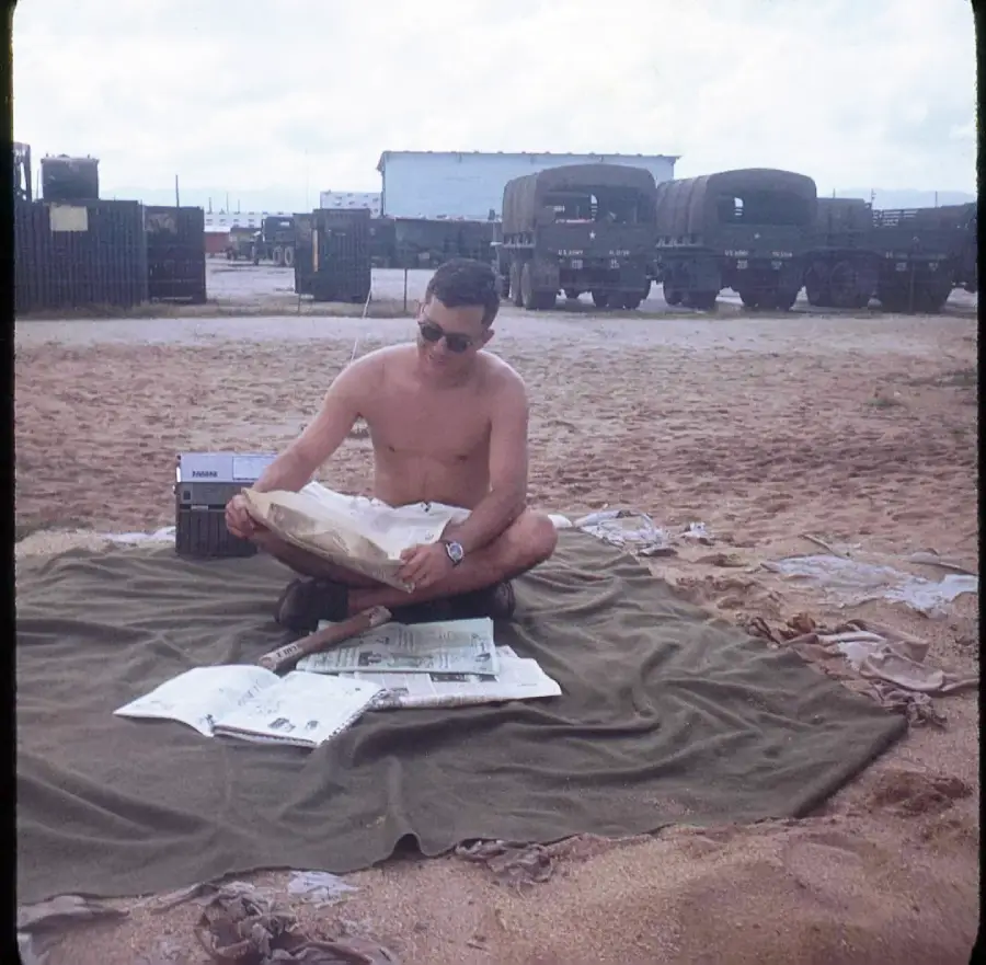 A shirtless man wearing sunglasses sits cross-legged on a blanket outdoors, reading a newspaper. Several papers and a radio are spread around him on the sand, with military trucks parked in the background.