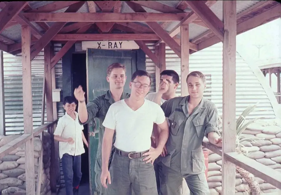 Four men in military attire stand and pose outside a building labeled "X-RAY"; one person in white stands in the background near the doorway. The building is surrounded by sandbags and has a corrugated metal exterior.