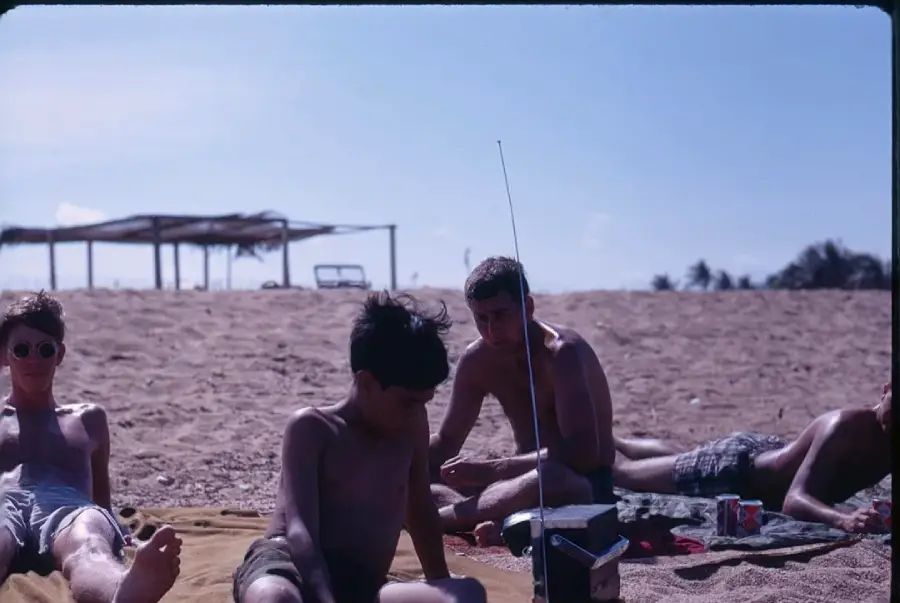Four young men sit and recline on a sandy beach under a clear sky, with a portable radio, a soda can, and towels laying nearby. A shaded structure and some trees are visible in the background.