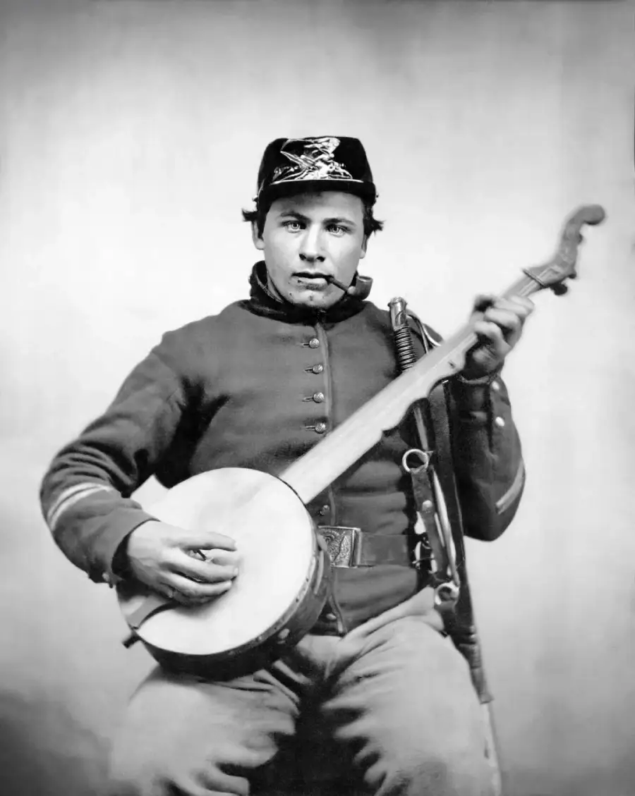 A black-and-white photo of a young Civil War-era soldier in uniform sitting and playing a banjo, looking directly at the camera with a neutral expression.
