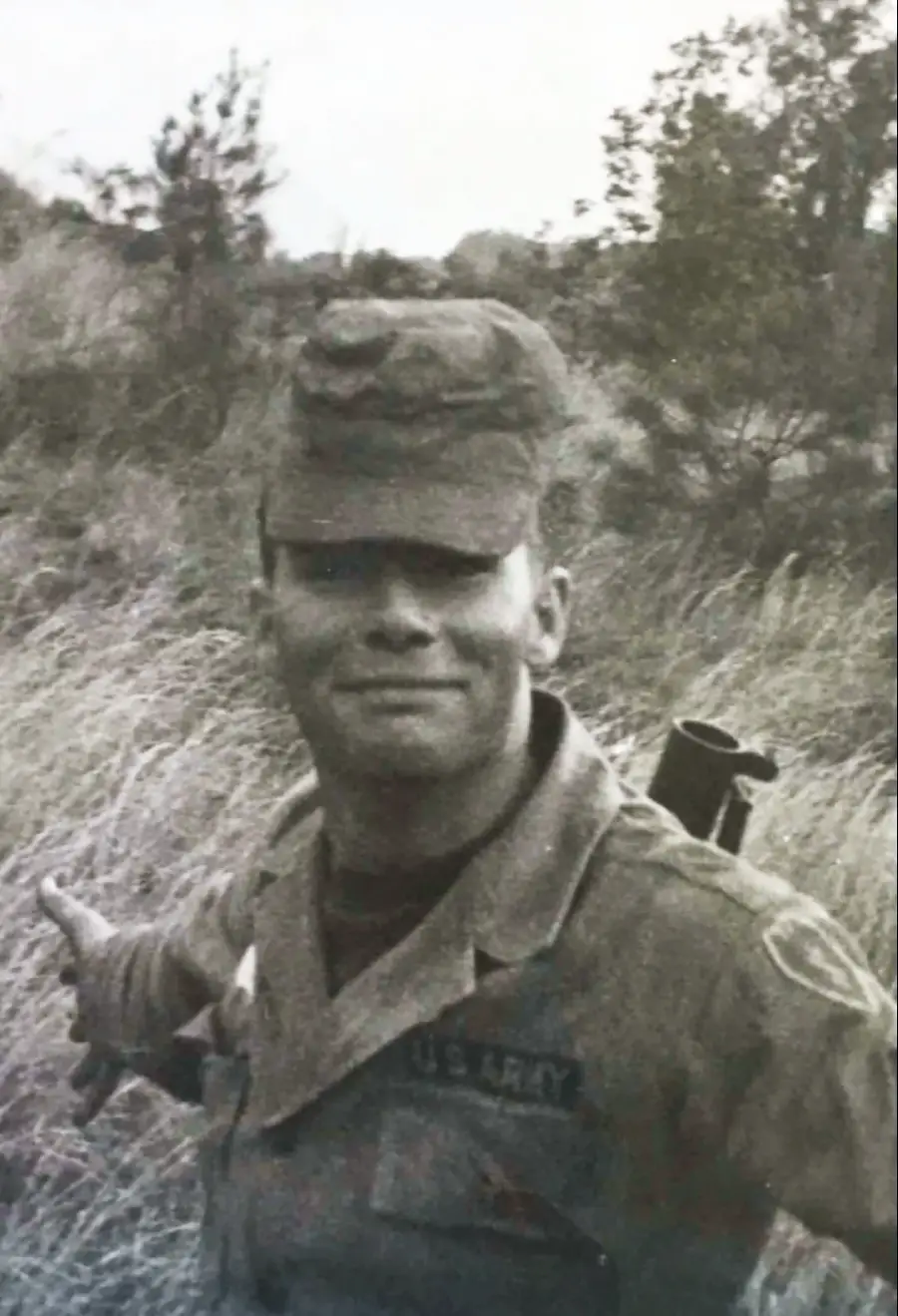 Black and white photo of a smiling U.S. Army soldier in uniform and cap, standing outdoors in tall grass with trees in the background. He holds one arm out to the side and has gear on his back.
