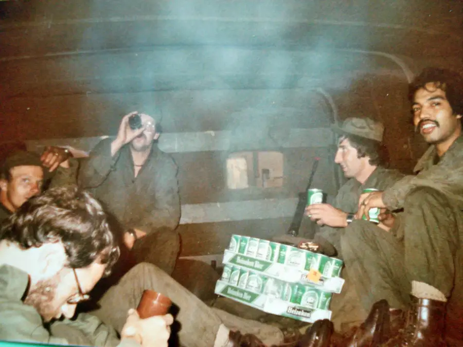 Five men in military uniforms sit together in a dimly lit room, some drinking from cans, with cases of Heineken beer on the floor between them. The atmosphere is relaxed and smoky.