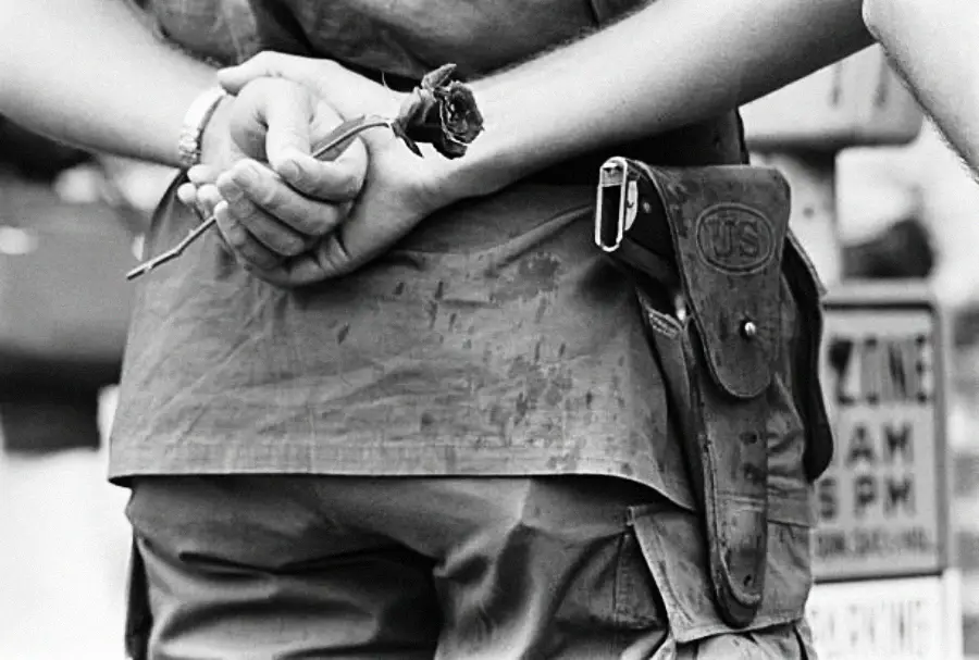 A person in a military or police uniform holds a wilted rose behind their back. The image is black and white, focusing on their hands and utility belt, with part of a street sign visible in the background.