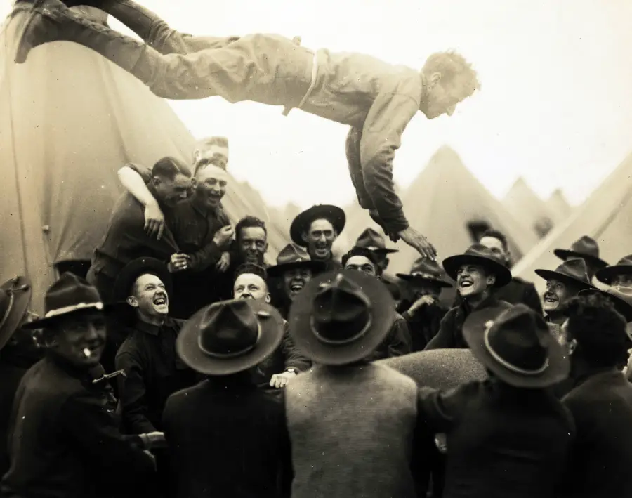 A group of soldiers in uniform laugh and smile as they toss a man high into the air above their heads, with military tents visible in the background.
