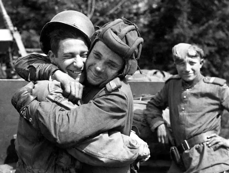 Two soldiers in uniform embrace and smile joyfully, while another soldier stands nearby, also smiling, with trees and military equipment in the background. The scene conveys camaraderie and relief.