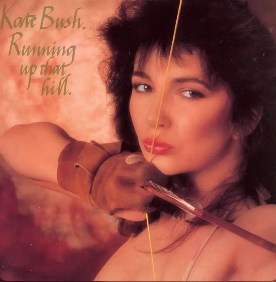 A woman holds a drawn bow and arrow close to her face. Text reads “Kate Bush. Running up that hill.” The background is a soft, warm brown, and the woman looks intently at the camera.