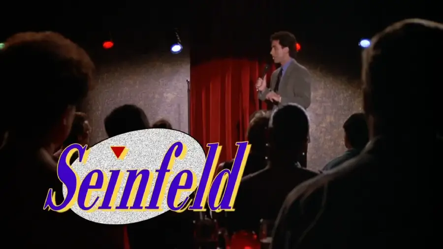 A man performs stand-up comedy on stage with red curtains, while an audience watches. The word "Seinfeld" appears in large yellow and purple letters across the image.