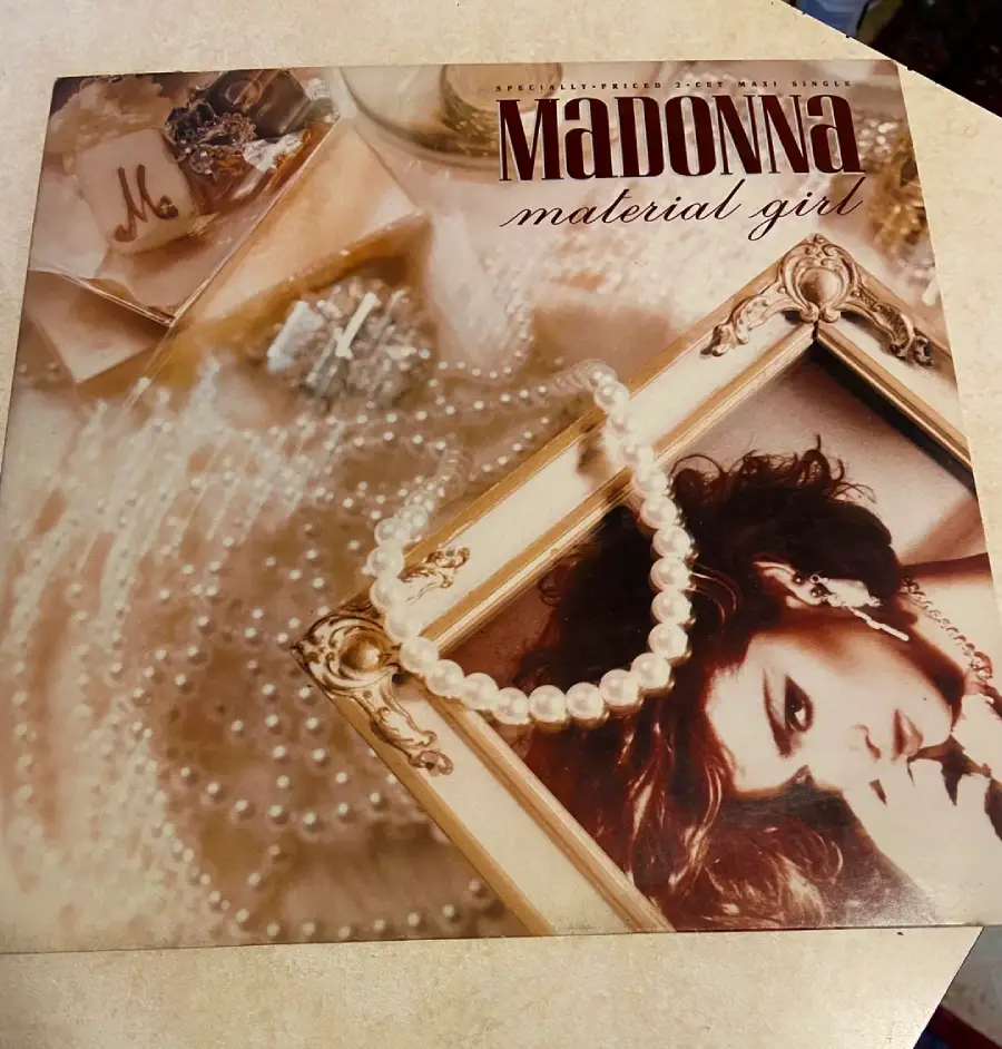 Material Girl album cover by Madonna, featuring a framed photo of Madonna, pearl necklaces, jewelry, and elegant decorations arranged on a cream-colored surface.