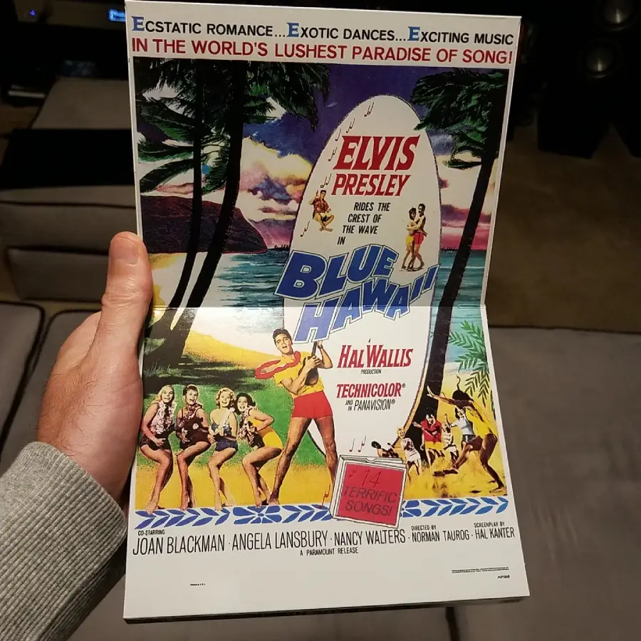 A hand holds a vintage "Blue Hawaii" movie poster featuring Elvis Presley, hula dancers, palm trees, and a beach scene. Bold, colorful text advertises the film’s stars and tropical setting.