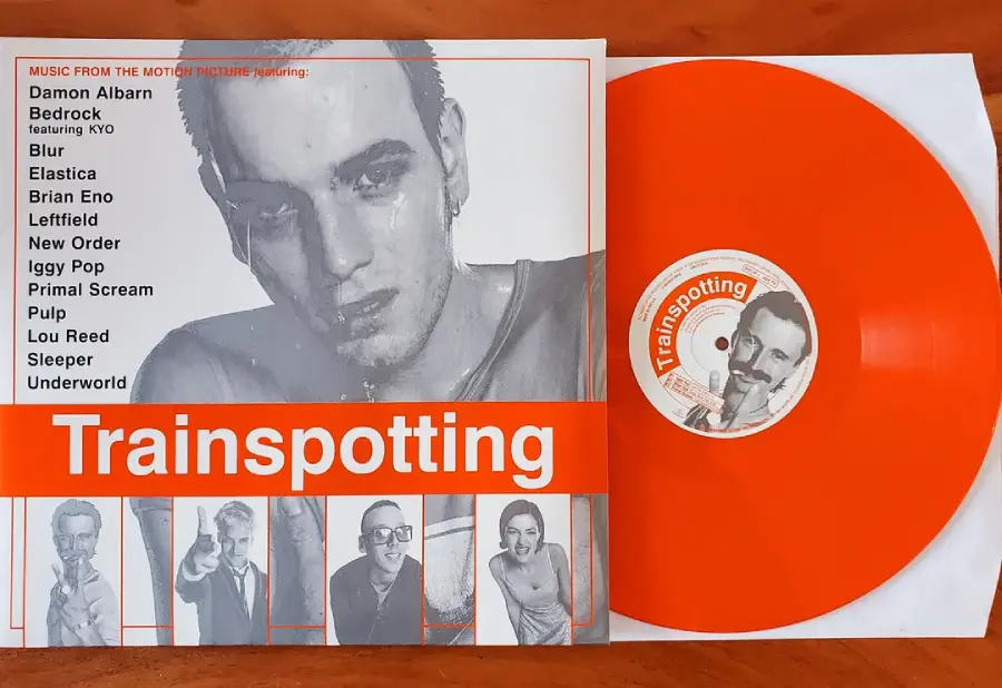 An orange vinyl record and its Trainspotting album cover featuring a grayscale photo of a man and a list of artists from the movie soundtrack. Small character photos line the bottom of the cover.