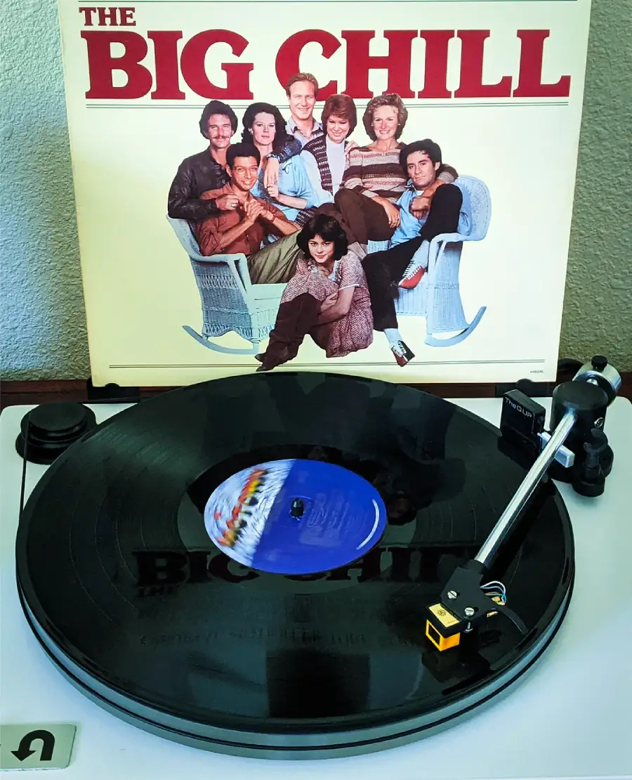 A vinyl record plays on a turntable in front of "The Big Chill" movie soundtrack album cover, which features a group of people sitting and smiling together.