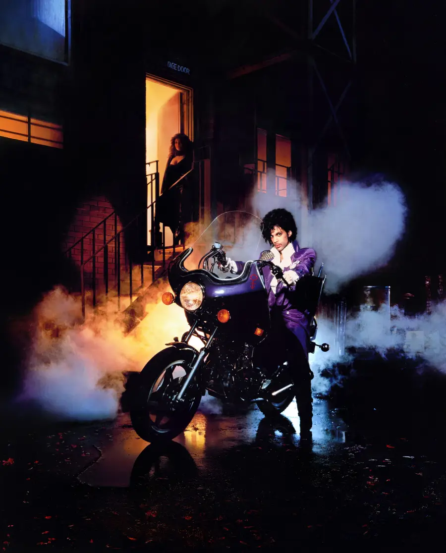 A person in a purple suit sits on a black motorcycle surrounded by smoke at night, with a woman standing in a lit doorway behind them on a set of stairs.