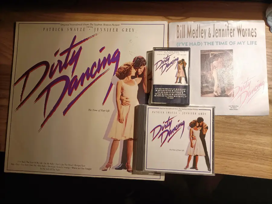 Various "Dirty Dancing" soundtrack formats, including a vinyl record, cassette, two CDs, and a 7-inch single, are displayed on a wooden surface. The cover art features a dancing couple in an embrace.
