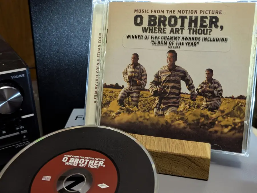 A CD and its case for "O Brother, Where Art Thou?" soundtrack are shown. The cover features three men in striped prison uniforms running in a field. Text claims it won five Grammy Awards, including "Album of the Year.