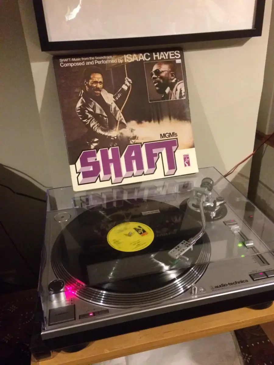 A vinyl record is playing on an Audio-Technica turntable. Behind it, the record sleeve for Isaac Hayes' "Shaft" soundtrack is displayed, showing Hayes with sunglasses and a guitar.