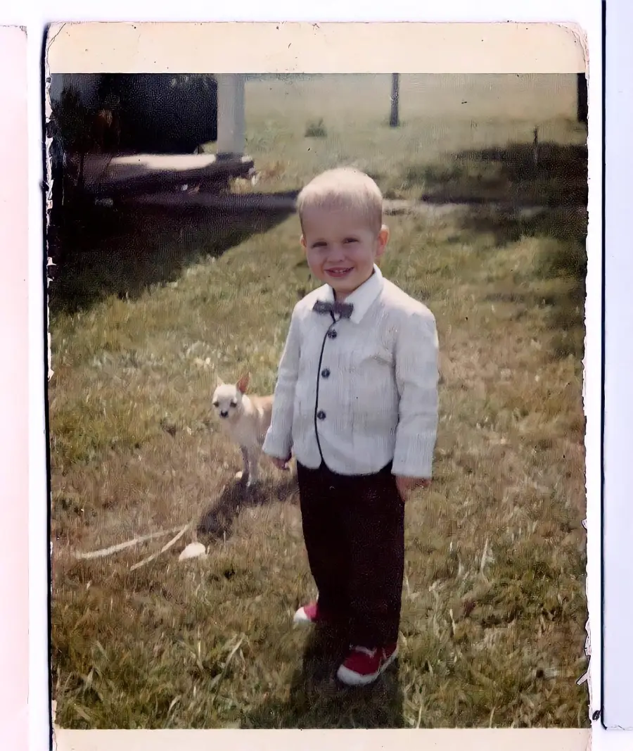 A young boy in a light-colored jacket, dark pants, and red shoes smiles while standing on grass. A small dog stands behind him. A porch and yard are visible in the background.