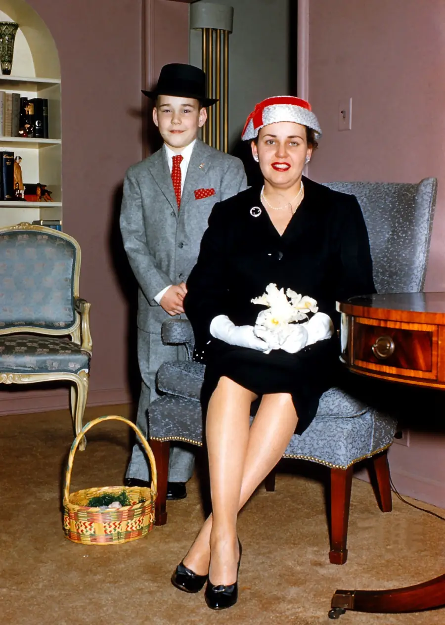 A woman in a black dress and red hat sits on a chair holding flowers, while a boy in a gray suit and black hat stands behind her. A basket sits on the floor nearby in a stylish, mid-century room.