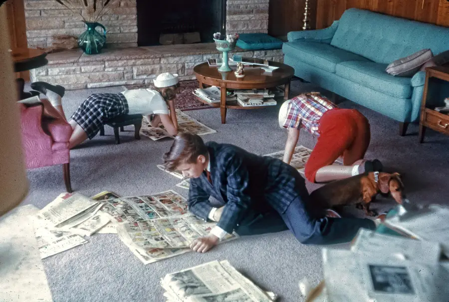 Three children kneel on a living room floor reading comic sections of newspapers, surrounded by scattered pages, while a dog lies nearby.