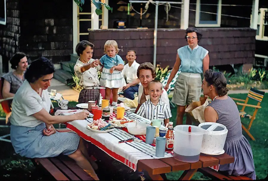 A group of women and children sit and stand around a picnic table outdoors, sharing food and drinks. The scene appears to be a casual summer gathering in a backyard.