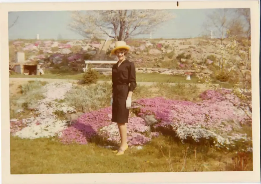 A woman in a yellow hat and black dress stands smiling in a garden with colorful flowers and green grass on a sunny day.