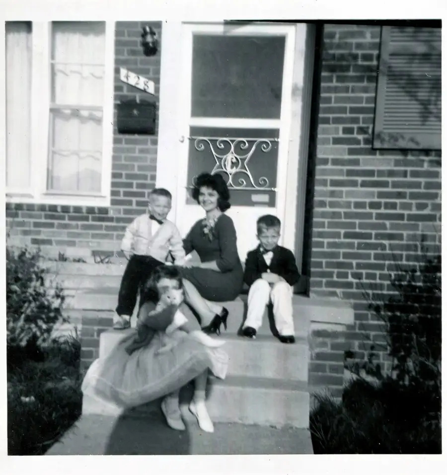 A woman and three children sit on the steps outside a brick house. The girl wears a dress, while the boys wear jackets. The group is smiling and posing for the photo.