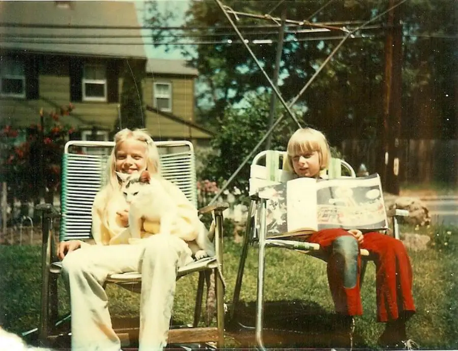 Two children sit outside on lawn chairs; one holds a white cat, the other reads a picture book. They are in a yard with a house in the background. The scene is sunny and relaxed.