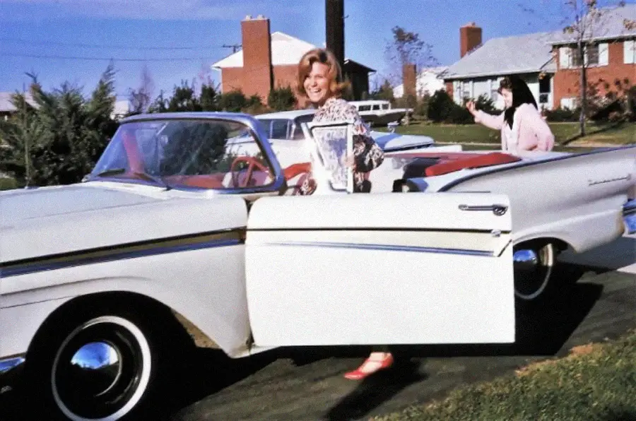 A woman stands by the open door of a white vintage convertible, smiling at the camera. Another person waves in the background near suburban houses on a sunny day.