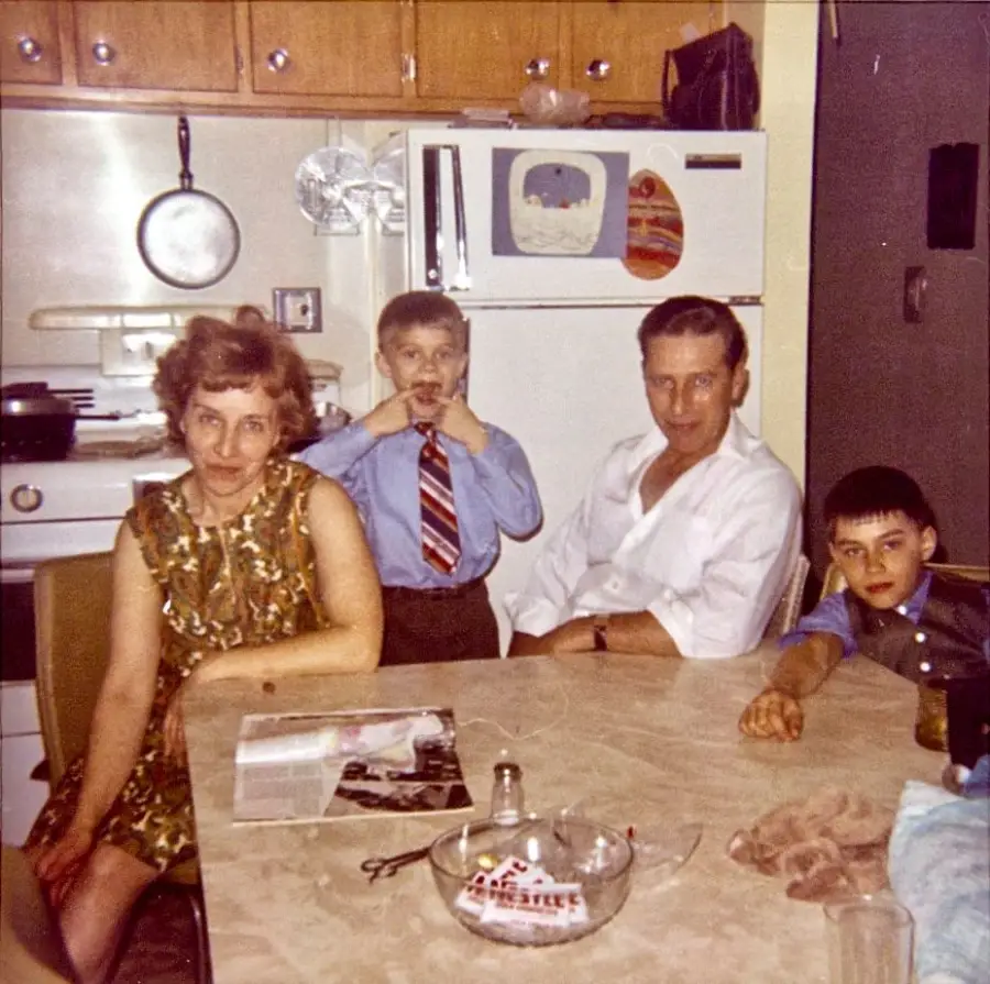 A woman, two boys, and a man sit at a kitchen table. One boy makes a funny face. The table holds a magazine and ashtray. The kitchen has wooden cabinets, a stove, and a refrigerator with magnets.