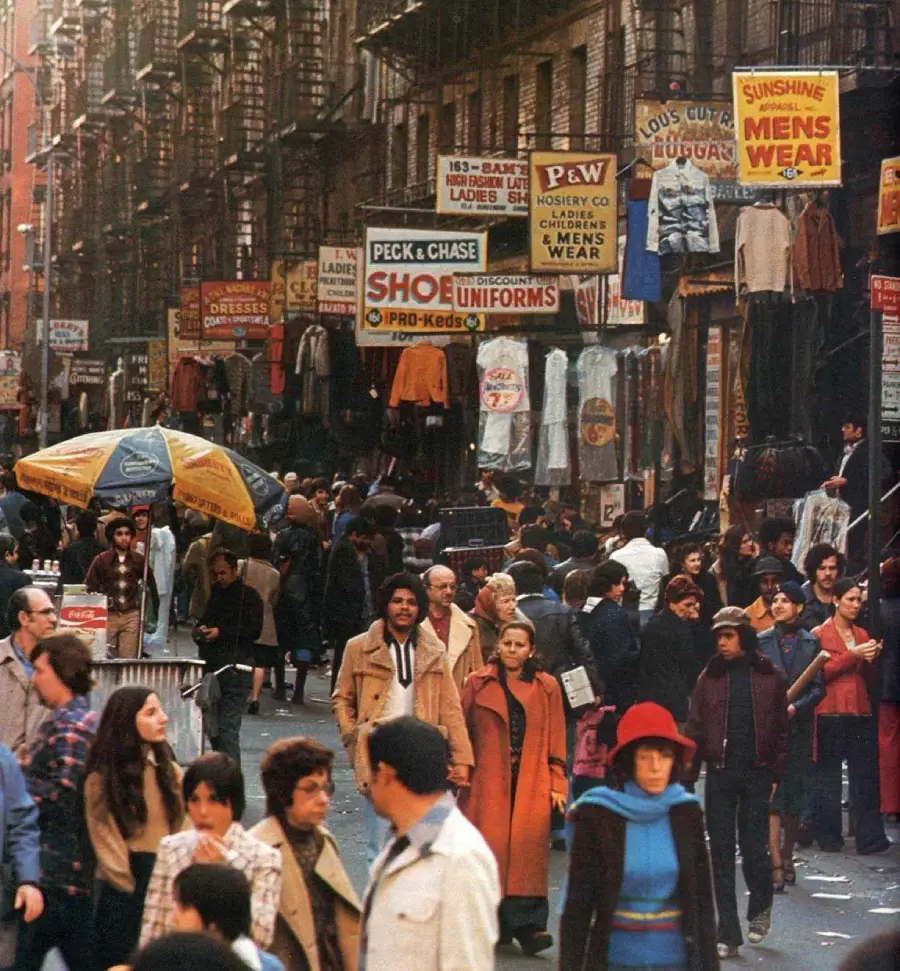 Crowded city street scene from the 1970s, with people walking among storefronts featuring colorful signs for shoes, uniforms, and clothing. Fire escapes line the buildings above.