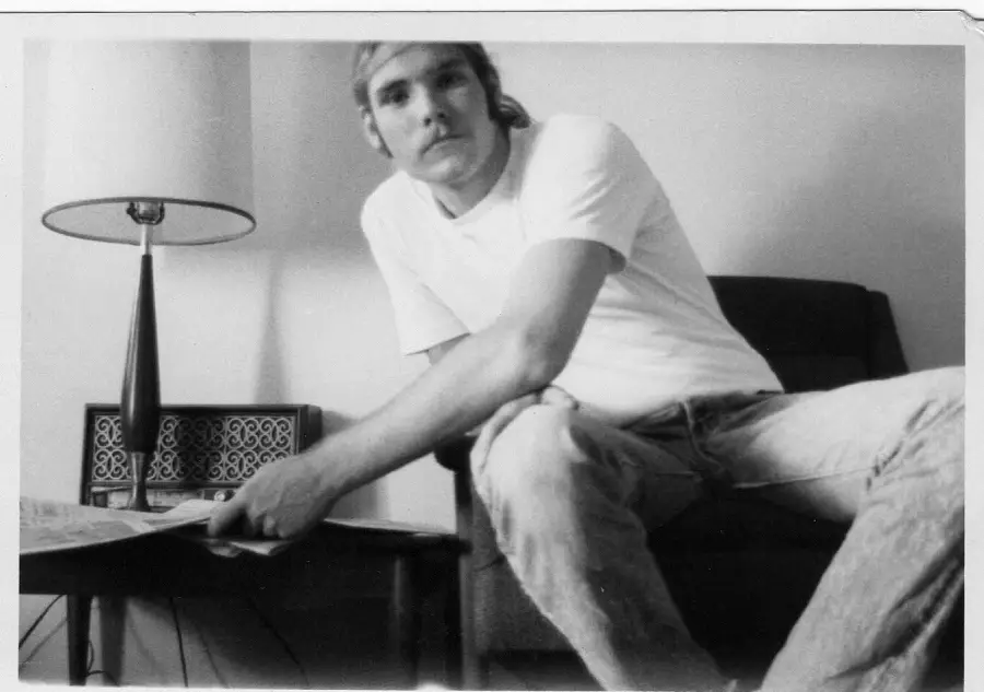A man in a white t-shirt and jeans sits on a chair next to a small table with a lamp, reaching toward a newspaper. The photo is black and white.