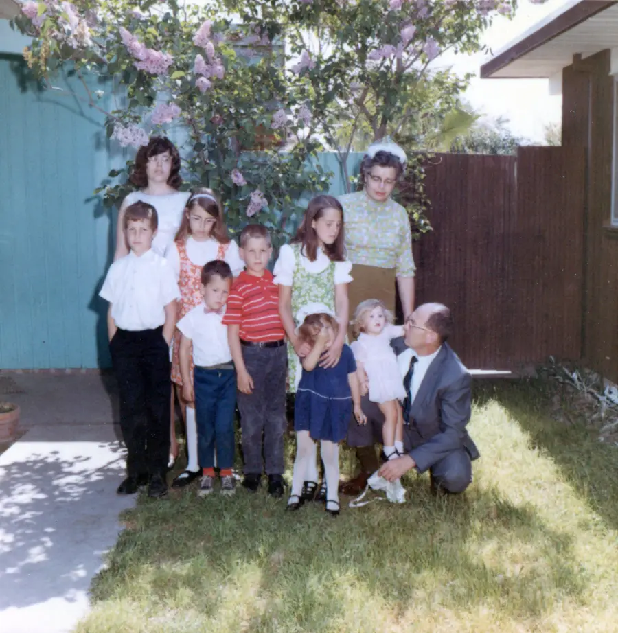 A family of nine poses in a sunny backyard. Seven children stand in front, with two adults—one woman standing, one man kneeling—beside them. There’s green grass and blooming flowers in the background.