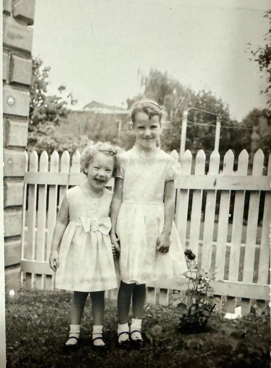 Two young girls in light-colored dresses stand outside in front of a white picket fence, smiling for the camera. A small plant is growing near their feet. The photo is black and white and appears vintage.