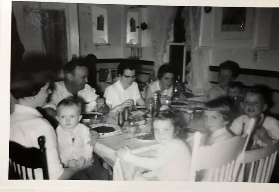A black and white photo shows a family, including adults and children, gathered around a dining table, sharing a meal in a cozy, vintage kitchen setting.