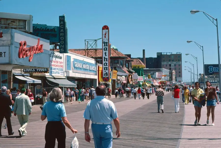 People walk along a sunny boardwalk lined with shops and restaurants, including a Nathan's Famous. The sky is clear and blue, and the boardwalk is busy with pedestrians enjoying the day.