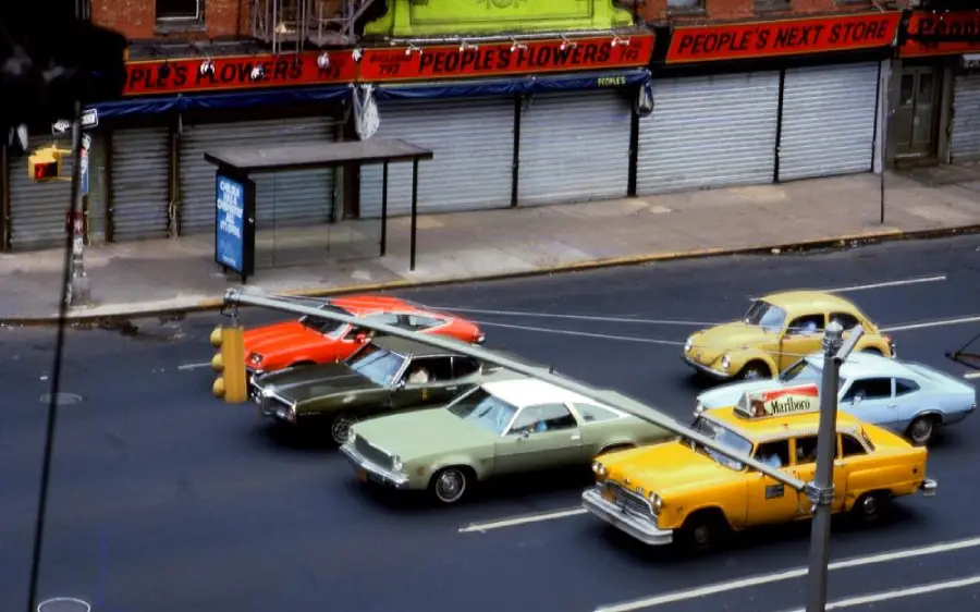 A row of vintage cars, including a yellow taxi and a yellow Volkswagen Beetle, stopped at a traffic light in front of shuttered shops on a city street.