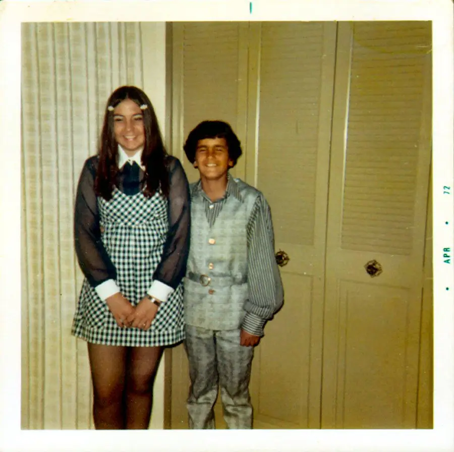 Two smiling children stand indoors; the girl wears a checkered dress with a white collar and hairclips, while the boy wears a denim vest and shirt. The background has beige closets. The photo is dated April 1972.