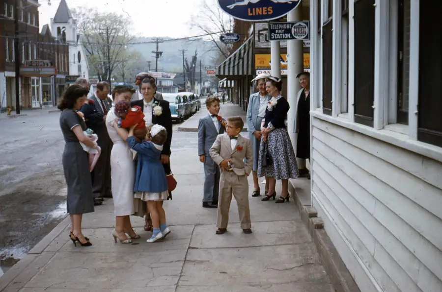 A group of people dressed in formal clothing stand on a small-town sidewalk, some talking while children stand together. A few women wear hats, and vintage cars and buildings line the quiet street in the background.